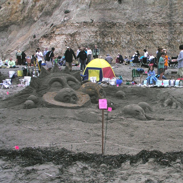 A sand sculpture of a cartoonish solar system with the suns arms wrapped around 8 planets.