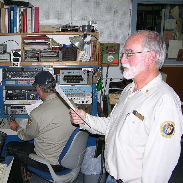 In an old radio station, a bearded man in a volunteer uniform reads aloud from a piece of paper.