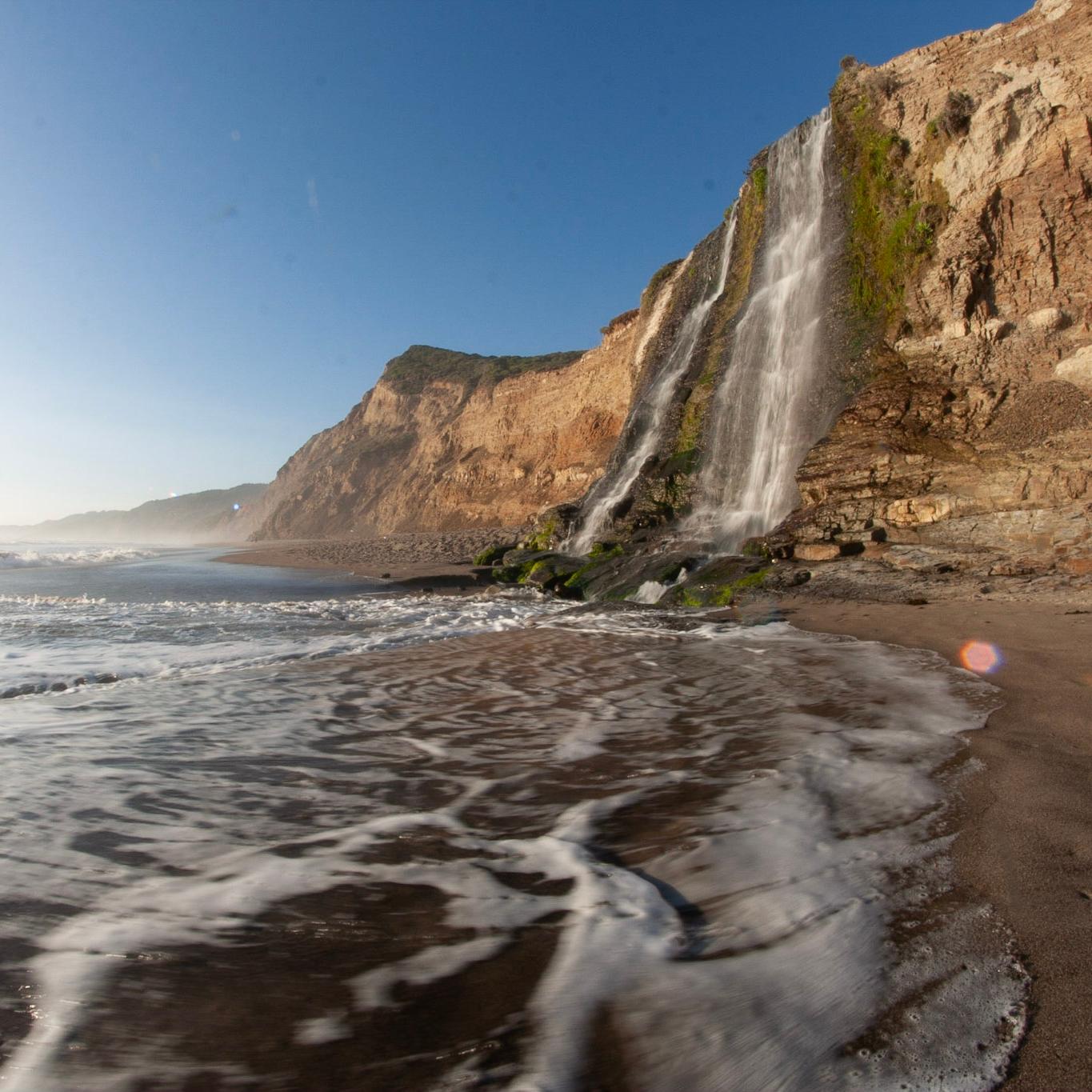 A 40-foot-tall waterfall cascades over coastal bluffs on the right onto a sandy ocean beach.