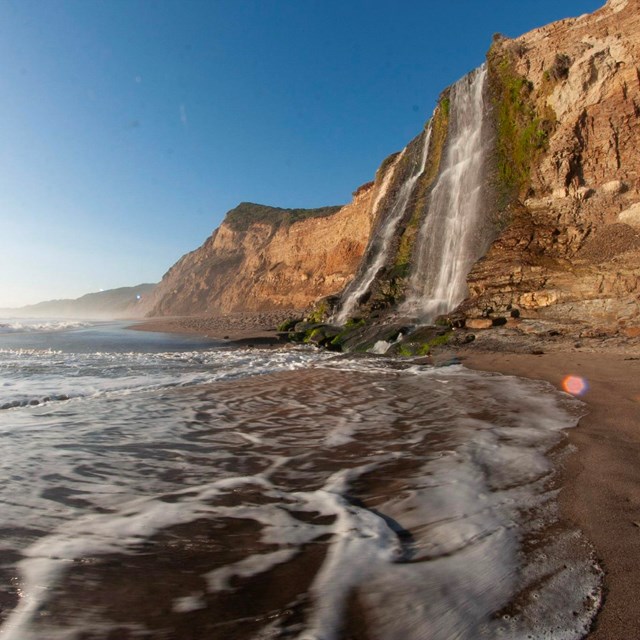 A 40-foot-tall waterfall cascades over coastal bluffs on the right onto a sandy ocean beach.