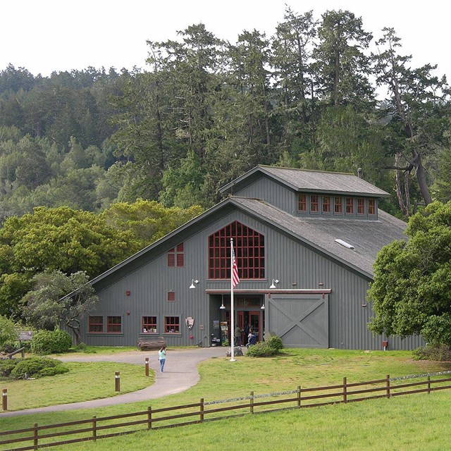A few visitors at a gray barn-like visitor center surrounded by fenced-pasture and trees.
