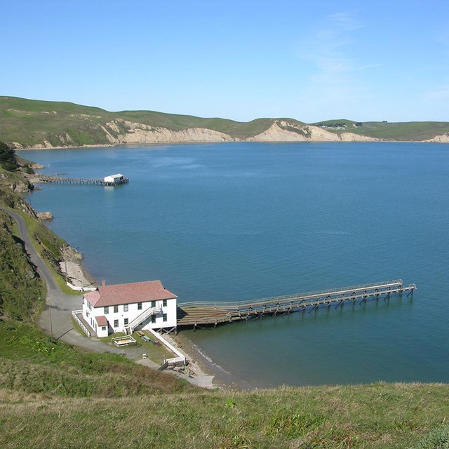 A two-story, white-sided, red-roofed building with a long dock at the edge of a blue bay.