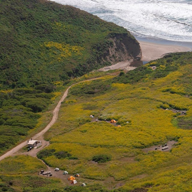An aerial photo of grassy campground with a few small tents and a dirt road near the beach. 