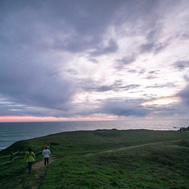 Two hikers walk along a dirt path atop a coastal bluff; a vast ocean and purple pink sunset behind. 