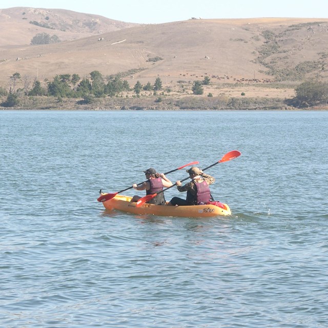 Two kayakers wearing purple PFDs paddling a yellow kayak on a calm bay.