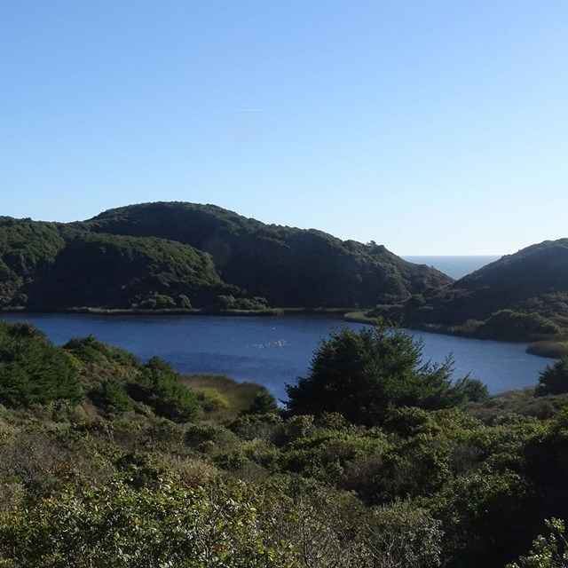 A small blue lake surrounded by brushy green hills with the ocean visible in the distance.