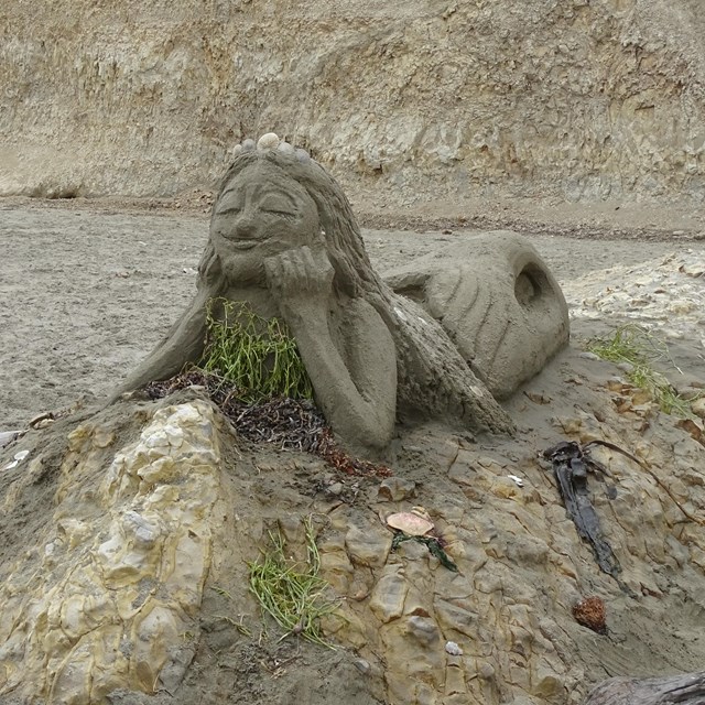 A medium-sized sand sculpture of a mermaid lying on her belly on top of a rock outcrop.
