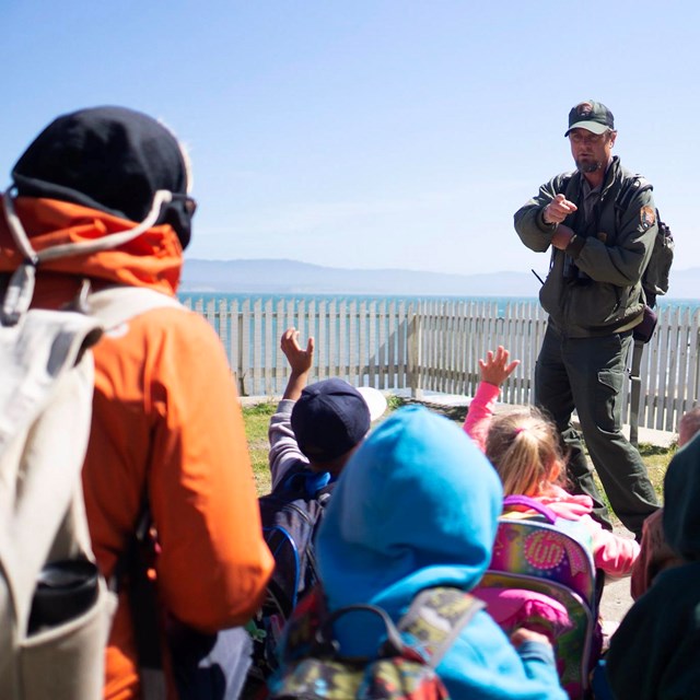 A park ranger wearing a ballcap speaks to a group of children near the ocean and seaside bluffs.