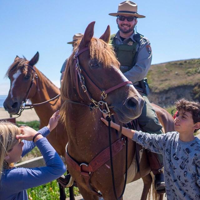 Two kids petting a horse atop which is a smiling park ranger.