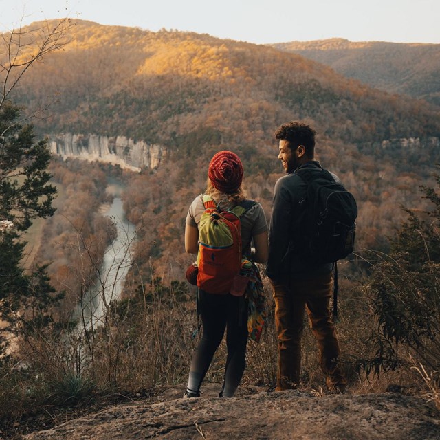 Two hikers standing on a rock outcrop high above a river that has cut a canyon through wood hills.