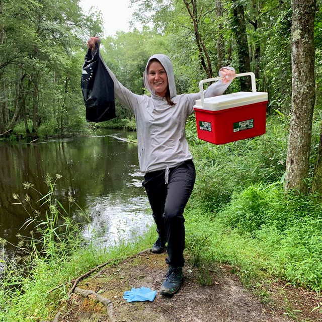 a smiling woman stands on a river bank holding up water quality sampling equipment