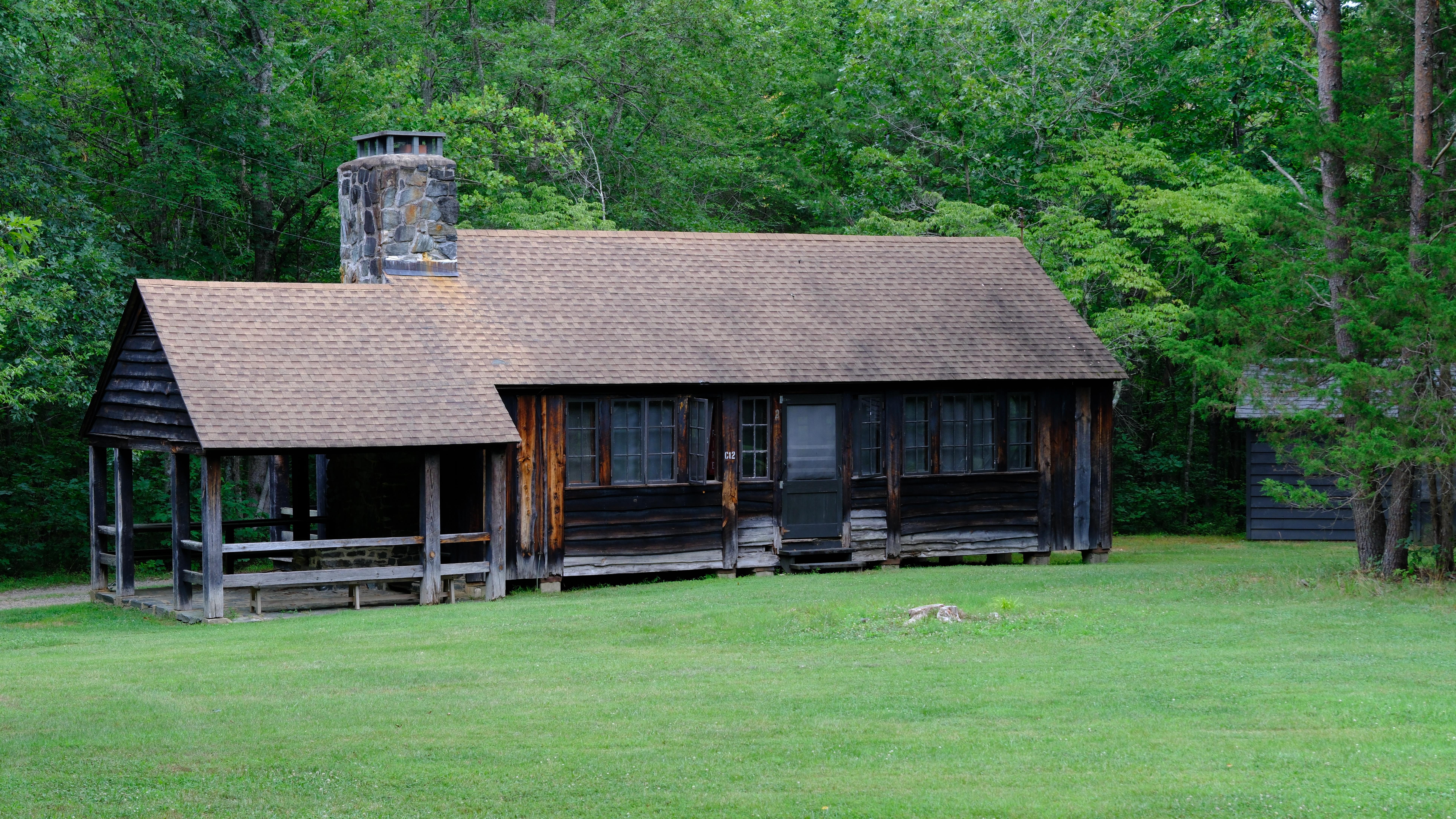 Wooden cabin with a stone porch and fireplace near a grassy meadow in a green forest