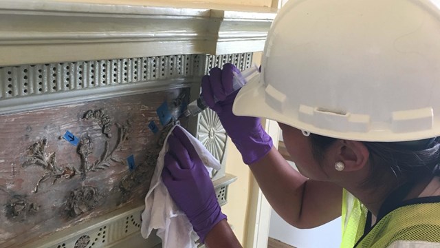 A conservator in hard hat and gloves applies glue to an intricate wooden mantel.