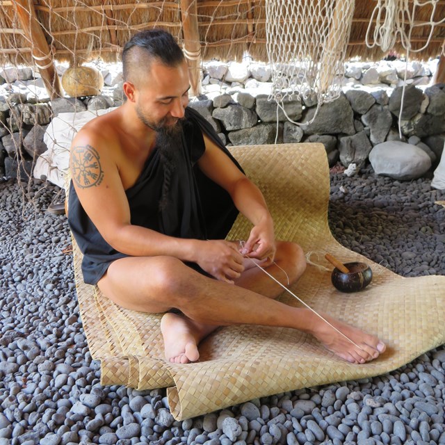 Man in traditional clothing practices traditional rope making while seated on a lauhala mat.