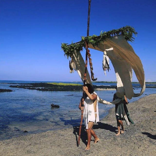 A man in white traditional clothing walks the shoreline holding the akua loa draped in white kapa.