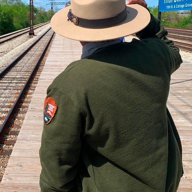 A ranger stands on a train platform looking away from the camera and instead at the tracks.