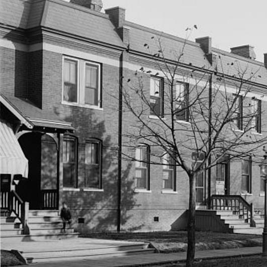 A black and white photo of newly built row houses in front of a dirt paved road.