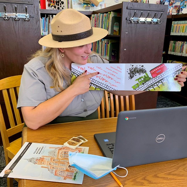 A ranger sits in a library holding up a book about trains to a webcam. A junior ranger book on table