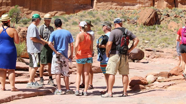 A group of people in summer clothes stands around a Park Ranger.