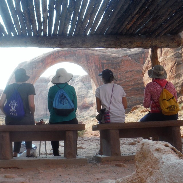 Four people sit under a wooden shade structure in the rain looking at Rainbow Bridge