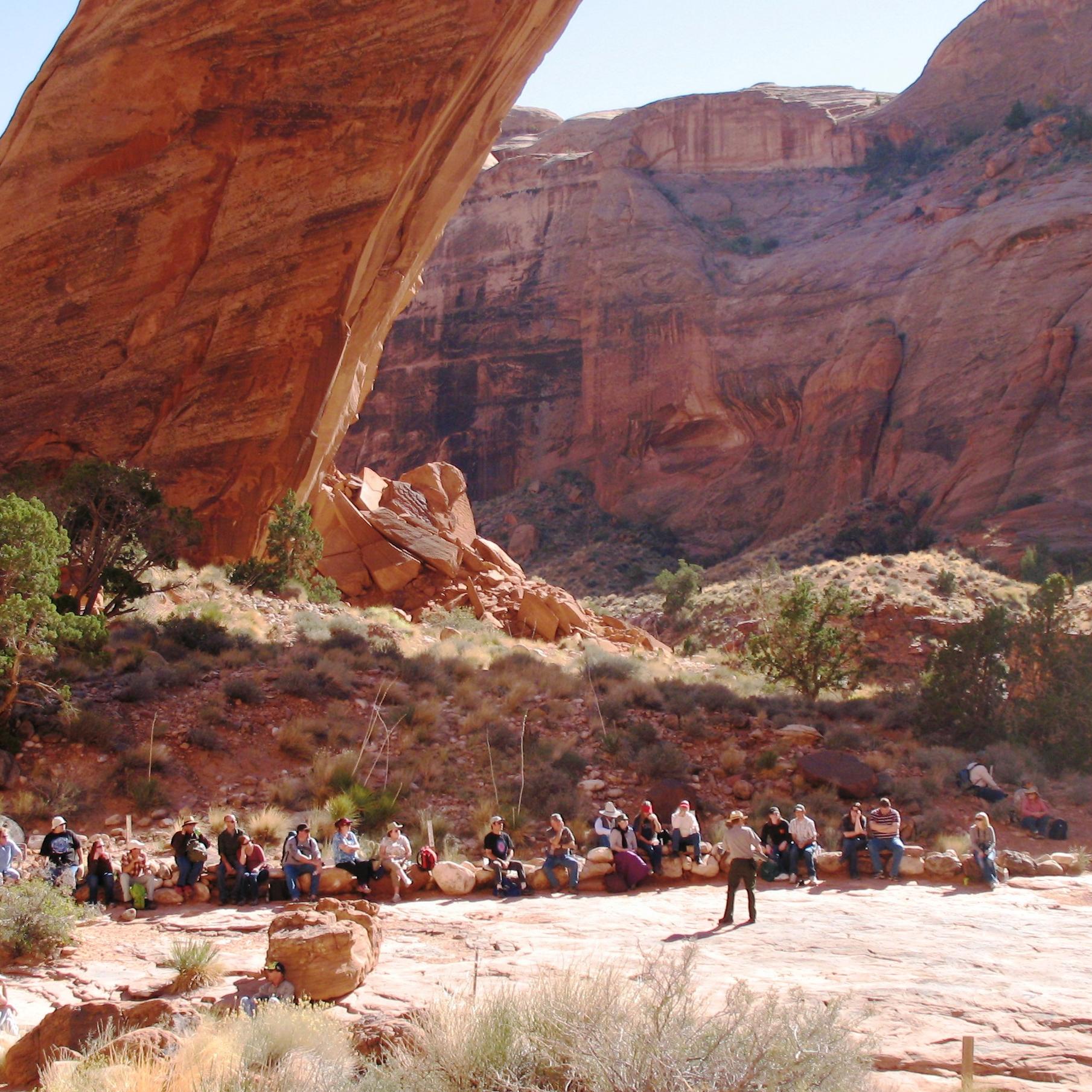 Perfect curved sandstone arch spans a small creek. A large group of people are gathered next to it.