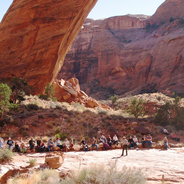 Perfect curved sandstone arch spans a small creek. A large group of people are gathered next to it.