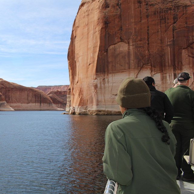 People standing at railing roof of a boat on calm waters. Sandstone cliffs surround the boat.