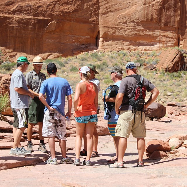 A group of people in summer clothes stands around a Park Ranger.