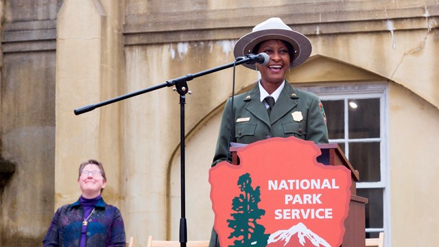 A ranger stands behind a podium with a National Park Service arrowhead.