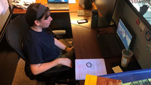 A young woman in a blue shirt talks to a group of people on a webcam at her desk