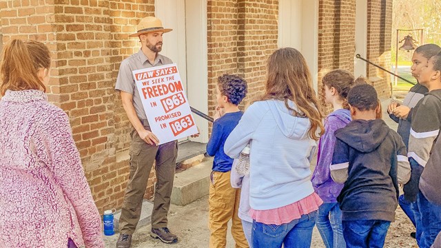 A park ranger holding a sign in front of a brick building, talking to a group of children. 