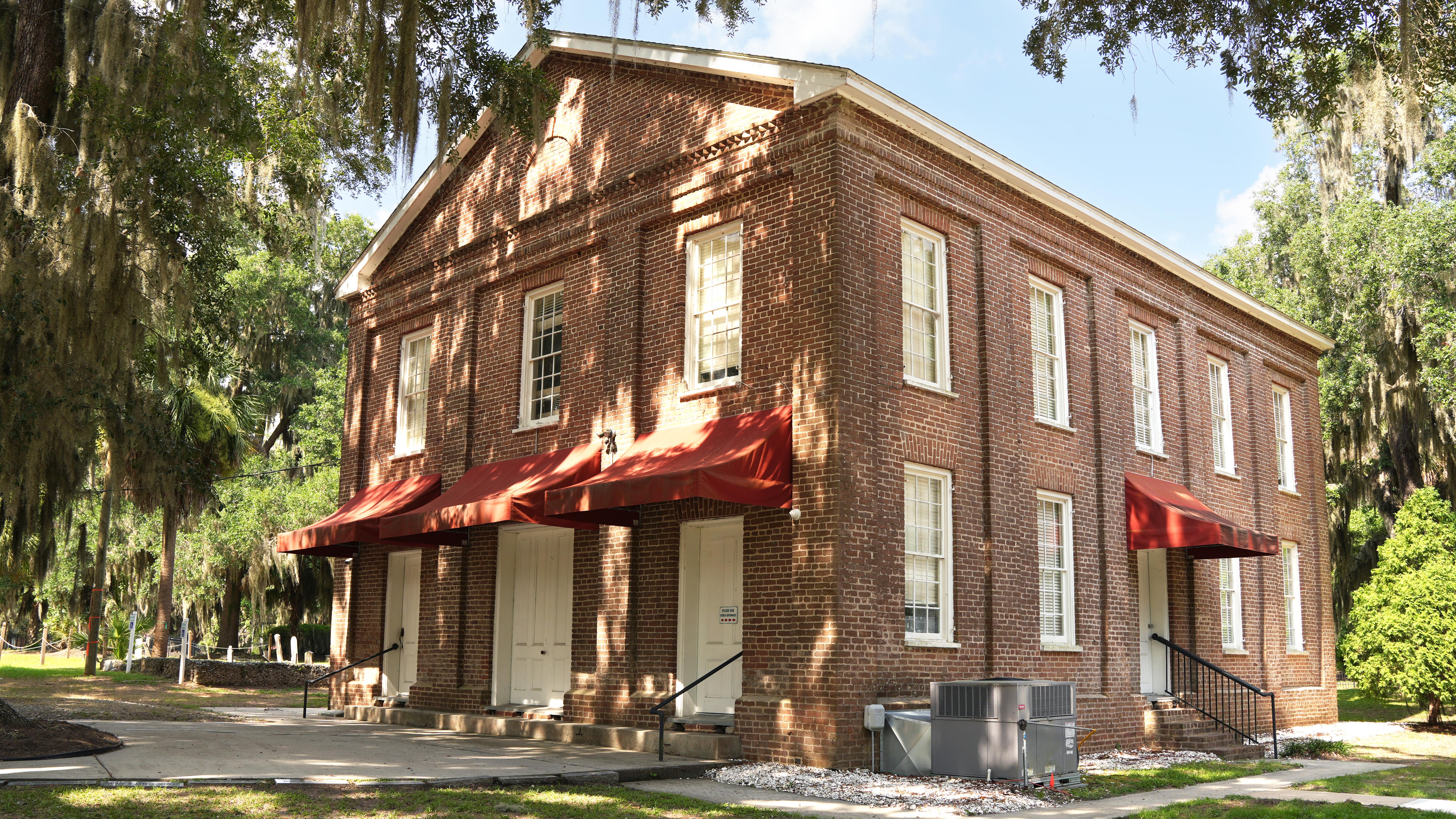 A two story brick church under oak trees and a blue sky.
