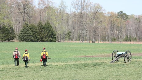 Three firefighters walking towards an open field and a cannon to their right.
