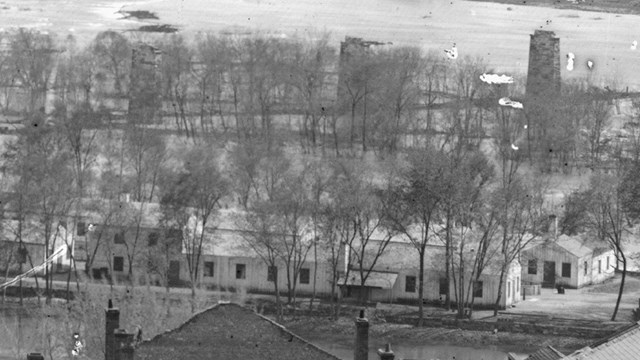 A black and white photograph of a group of small, white buildings on an island in a river.