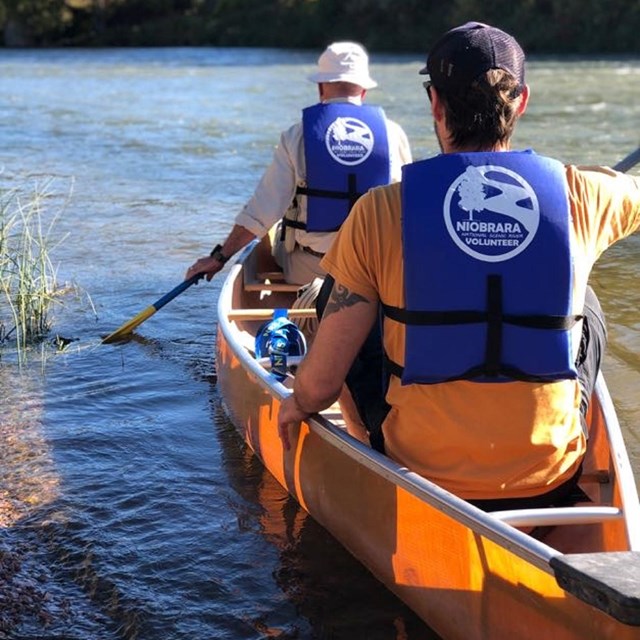 Two people in blue lifejackets launch a canoe into a calm river