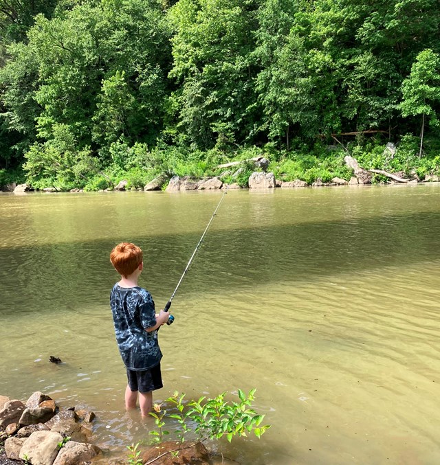 A young boy holds a fishing rod while standing on the edge of a murky river.