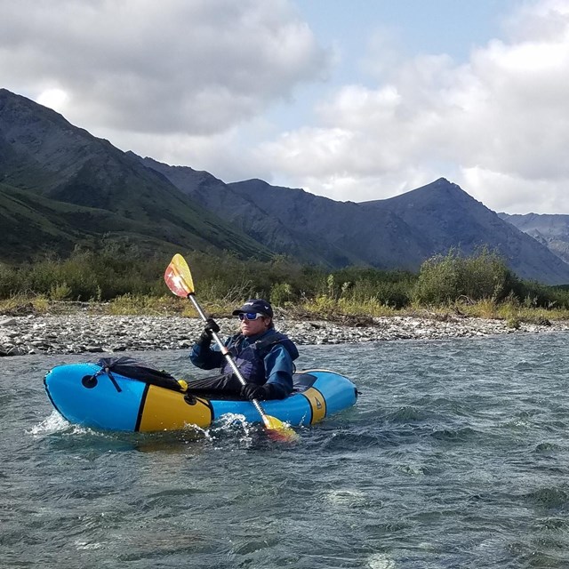 A person in an inflatable kayak paddles down a river with mountains in the background.