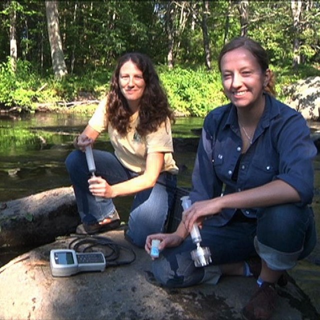 Two scientists hold water quality measuring equipment while sitting on rocks in a river 