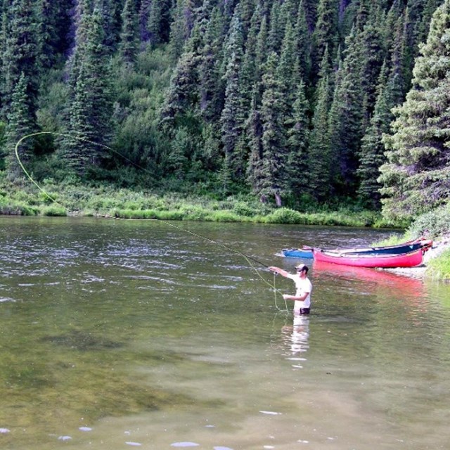 A person stands in the flowing water of a river fly fishing with a red canoe on the bank