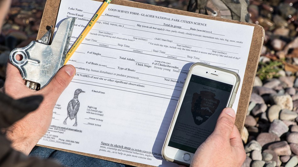 A hand holds a clipboard with a data sheet, a pencil, and a smartphone