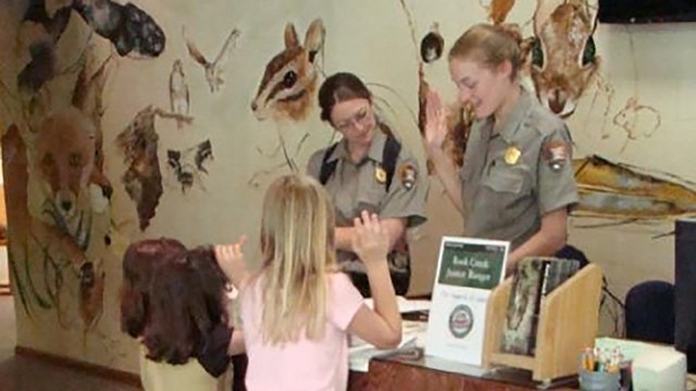 Two female rangers stand behind a desk. Three young girls look over the edge of the desk at them