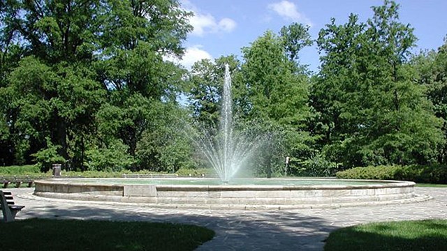 A low stone wall encloses a round pool with a fountain spurting up from the middle