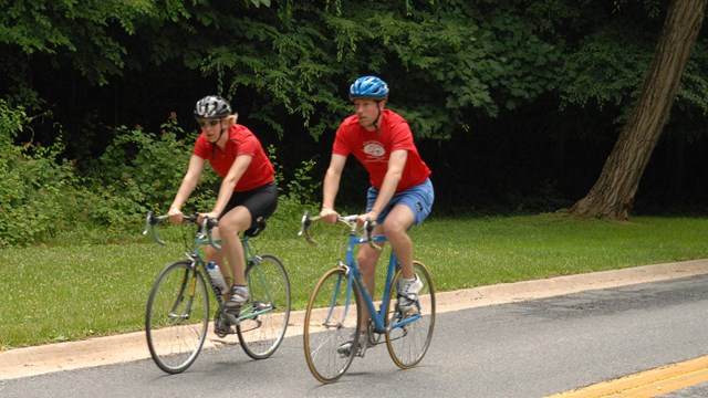 Two bicyclists ride next to each other wearing matching red shirts 