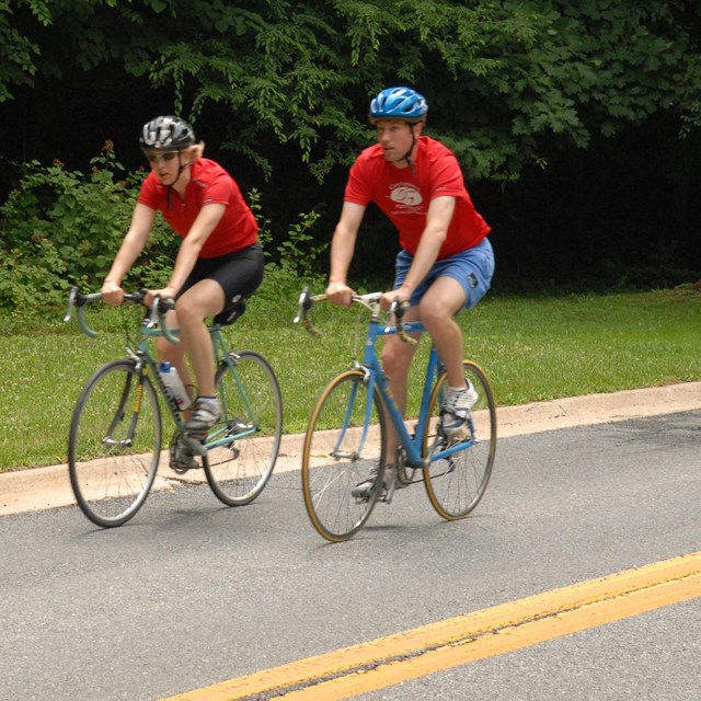 Two bicyclists ride next to each other wearing matching red shirts 