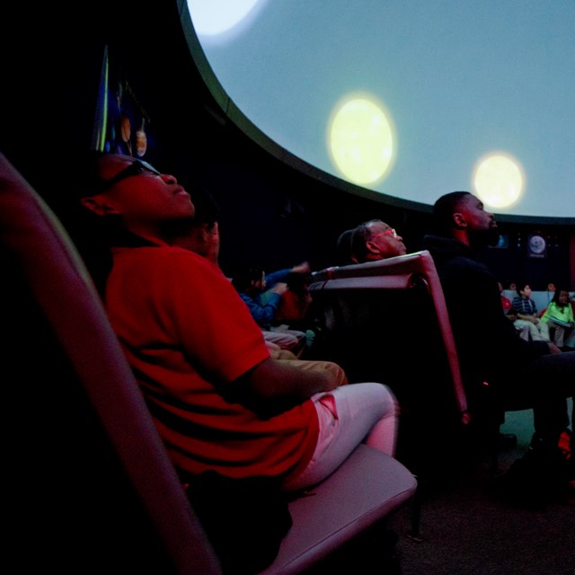 A child sitting with a crowd of people stares up at at the planetarium ceiling during a program. 