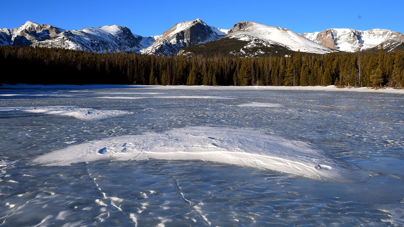 View of Bierstadt Lake in winter, frozen over and dusted with snow. Mountain peaks in the distance