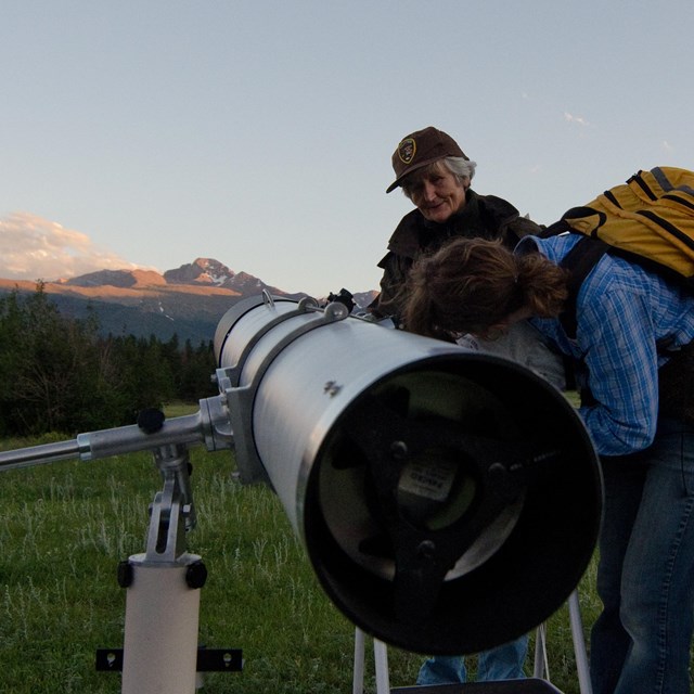 An NPS volunteer is standing by a telescope. A visitor is looking in the telescope