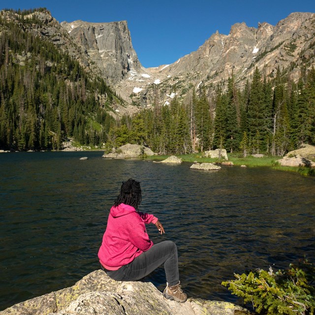 A person is sitting on a rock at the shore of an alpine lake, looking out at the lake