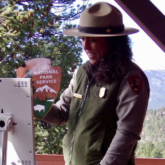 A ranger holds up the NPS arrowhead emblem in front of an iPad with a mountain backdrop