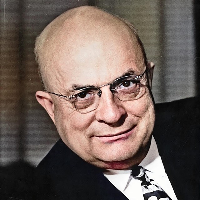 A historic photo of a bald older man wearing glasses and a suit. 